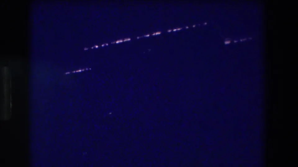 8mm home movie frame, Acapulco Mexico 1982 — two separate bands of elevated lights visible over dark coastal water in a deeply blue-cast, grainy frame.
