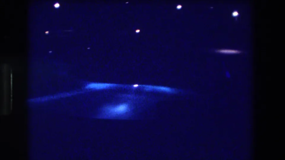 Night pan on 8mm film, Acapulco Mexico 1982—a diffuse glowing area, possibly a lit pool or courtyard, surrounded by deep blue darkness and scattered distant lights.