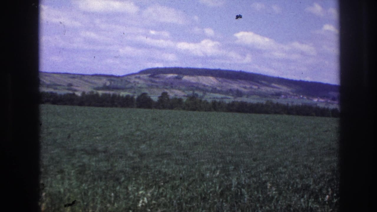 8mm home-movie frame from France, c.1967 — broad green agricultural field with treeline and distant rolling hills under a partly cloudy blue sky.