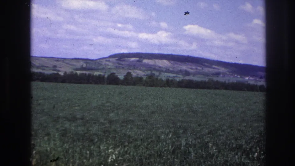 8mm home-movie frame from France, c.1967 — broad green agricultural field with treeline and distant rolling hills under a partly cloudy blue sky.