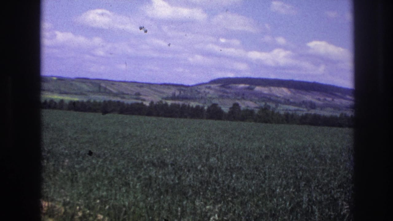 Agricultural crop field and rolling hills under a partly cloudy blue sky, 8mm film with visible grain and dust specks.