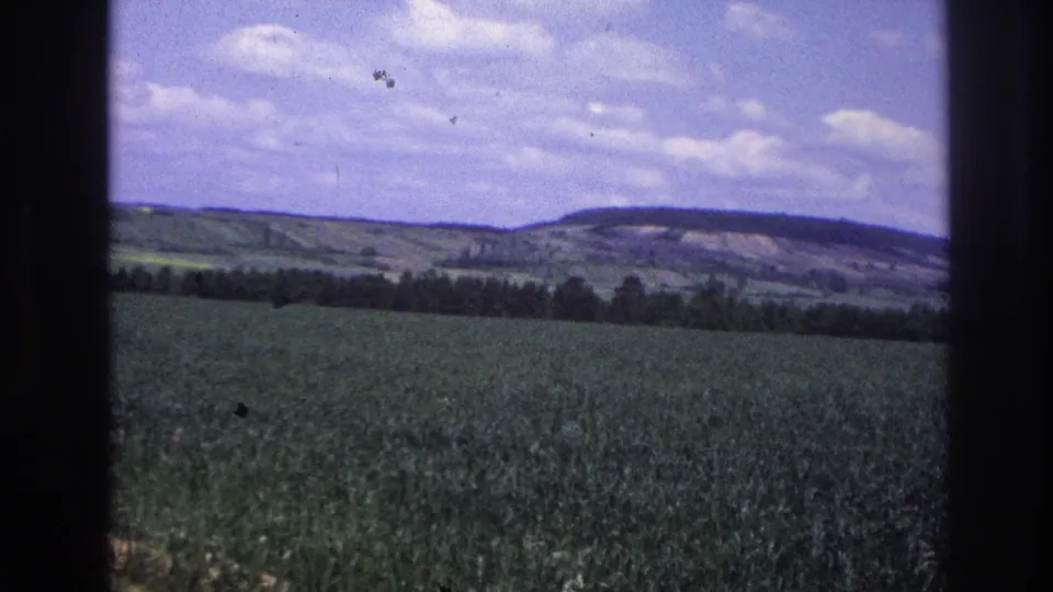 8mm home-movie frame from France, c.1967 — green crop field, dark treeline, and distant hills under partly cloudy sky; slight camera motion from moving vehicle.