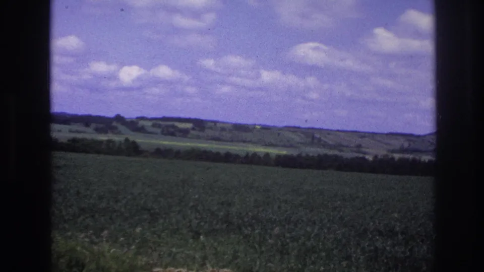 Final 8mm home-movie frame panning across French countryside, c.1967 — crop field and rolling hills under expansive blue sky with white cumulus clouds.