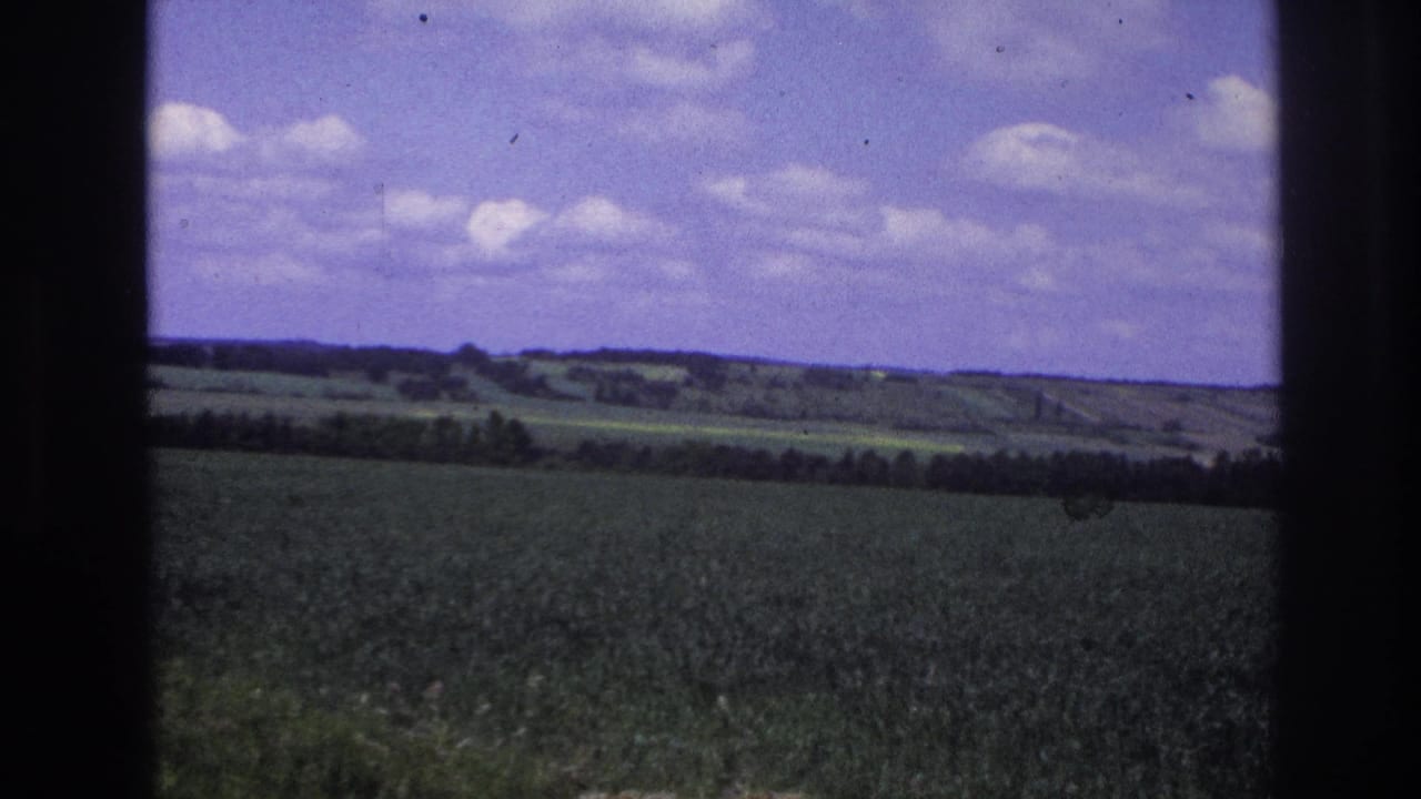 8mm film frame from France, 1967: a cultivated green field stretches to a wooded horizon under a blue sky with white clouds, shot through a vehicle window.