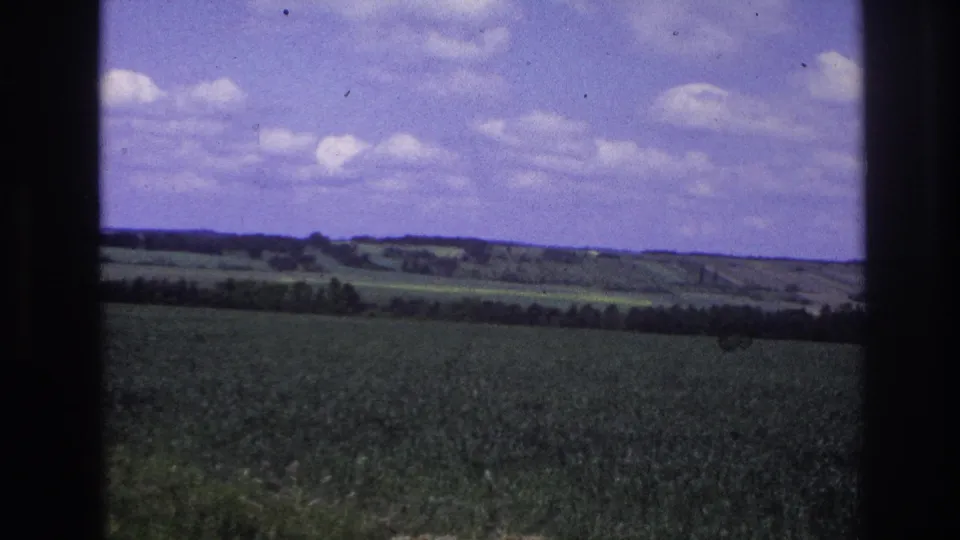 8mm film frame from France, 1967: a cultivated green field stretches to a wooded horizon under a blue sky with white clouds, shot through a vehicle window.