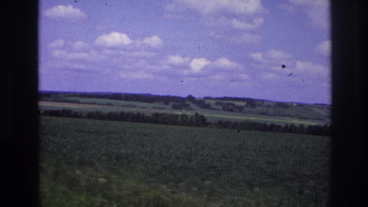8mm travel footage, France 1967: green cultivated field and wooded hills under a lavender-tinted sky, shot through a moving vehicle window.