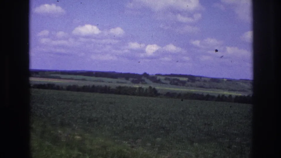 8mm travel footage, France 1967: green cultivated field and wooded hills under a lavender-tinted sky, shot through a moving vehicle window.