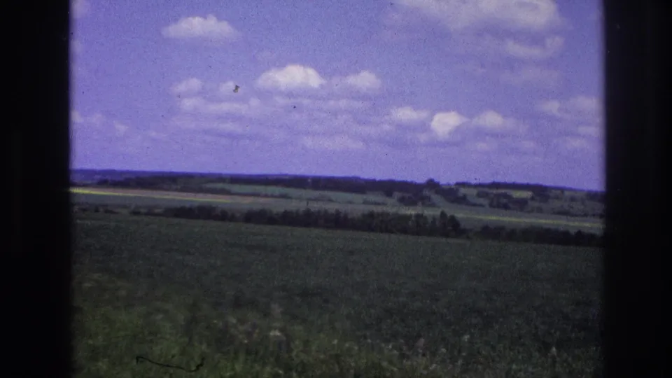 8mm footage, France 1967: a broad green field leads to distant hills with a possible bright crop strip on the horizon, framed by a vehicle window under cumulus clouds.
