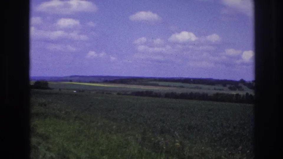 Final 8mm frame, France 1967: expansive green countryside and distant tree-lined hills under a purple-tinted sky with white cumulus clouds.