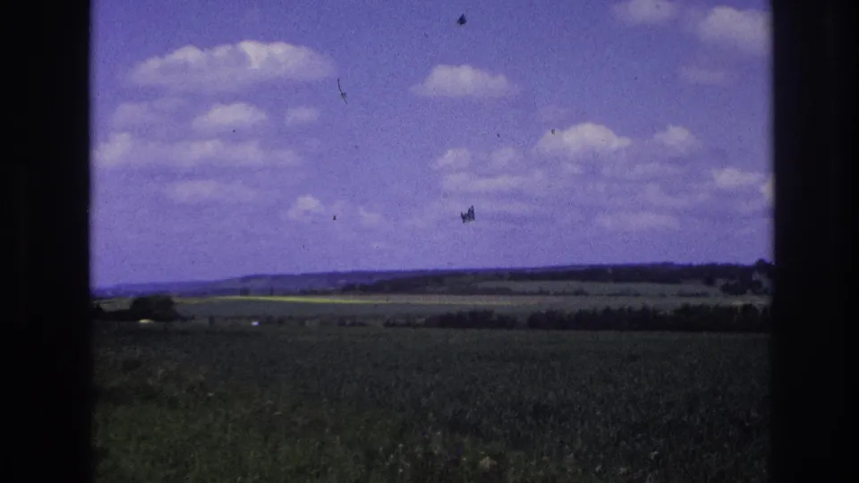 1967 France home-movie frame showing open green fields, distant tree lines, low hills, and a clouded blue sky from a moving-vehicle viewpoint.