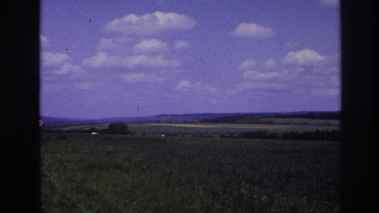 1967 France frame with farmland, distant hills, a low tree line, and scattered white clouds viewed from the road.