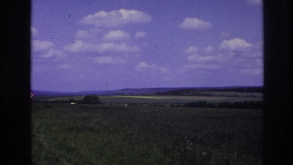 1967 France frame with farmland, distant hills, a low tree line, and scattered white clouds viewed from the road.