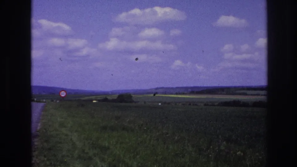1967 France moving-vehicle frame showing a paved rural road, grassy verge, green fields, a red-bordered traffic sign, and low hills.