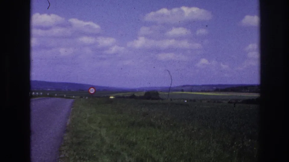 1967 France countryside frame showing the road edge, green fields, a red-bordered circular roadside sign, distant hills, and a bright clouded sky.