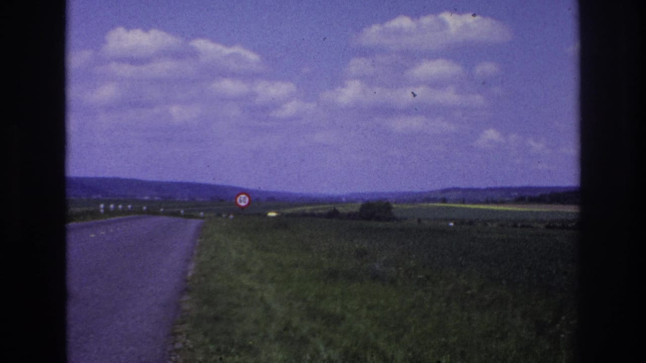 1967 France rural road seen from a moving vehicle, with green fields, distant hills, cloud cover, and a red-bordered 50 speed limit sign ahead.