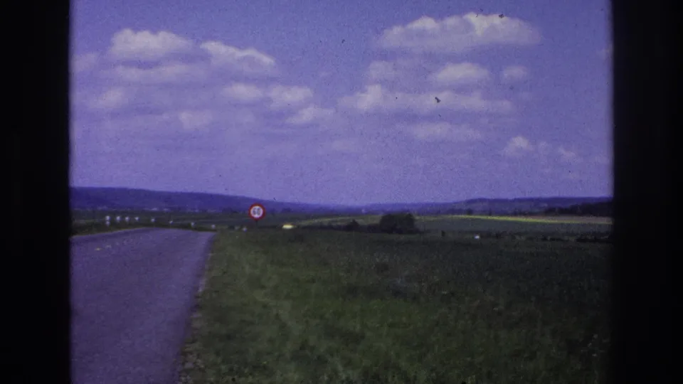 1967 France rural road seen from a moving vehicle, with green fields, distant hills, cloud cover, and a red-bordered 50 speed limit sign ahead.