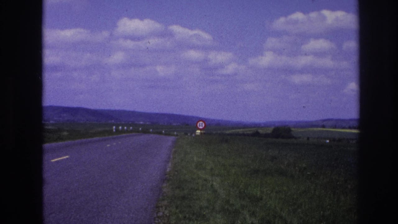 French countryside road in 1967 with grassy verges, low hills, roadside marker posts, and a 50 speed limit sign visible near the right shoulder.