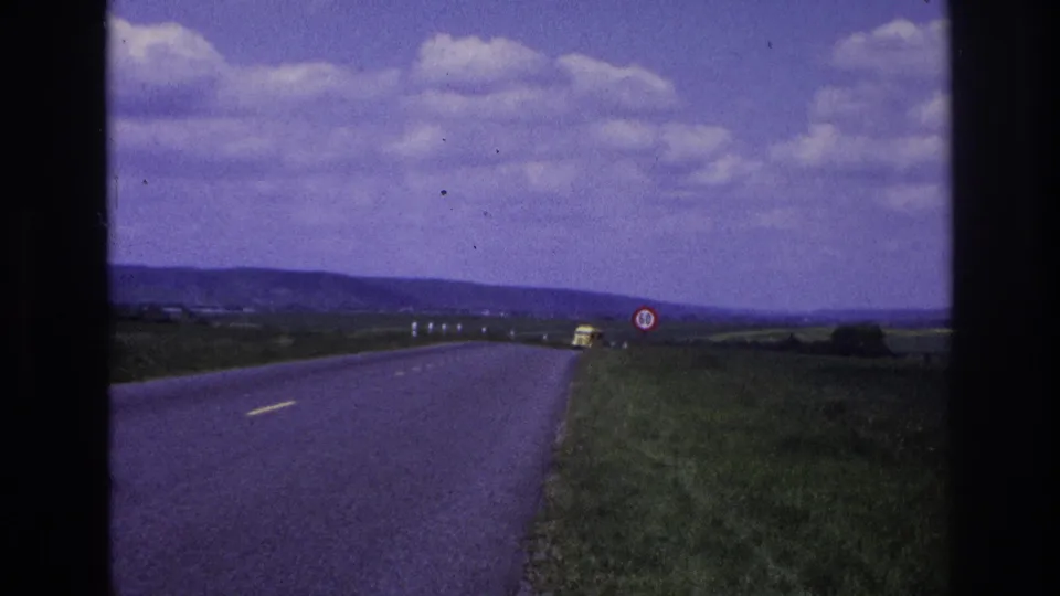 A small yellow car approaches in the distance on a 1967 French rural road, with a 50 speed limit sign, fields, hills, and a clouded sky around it.
