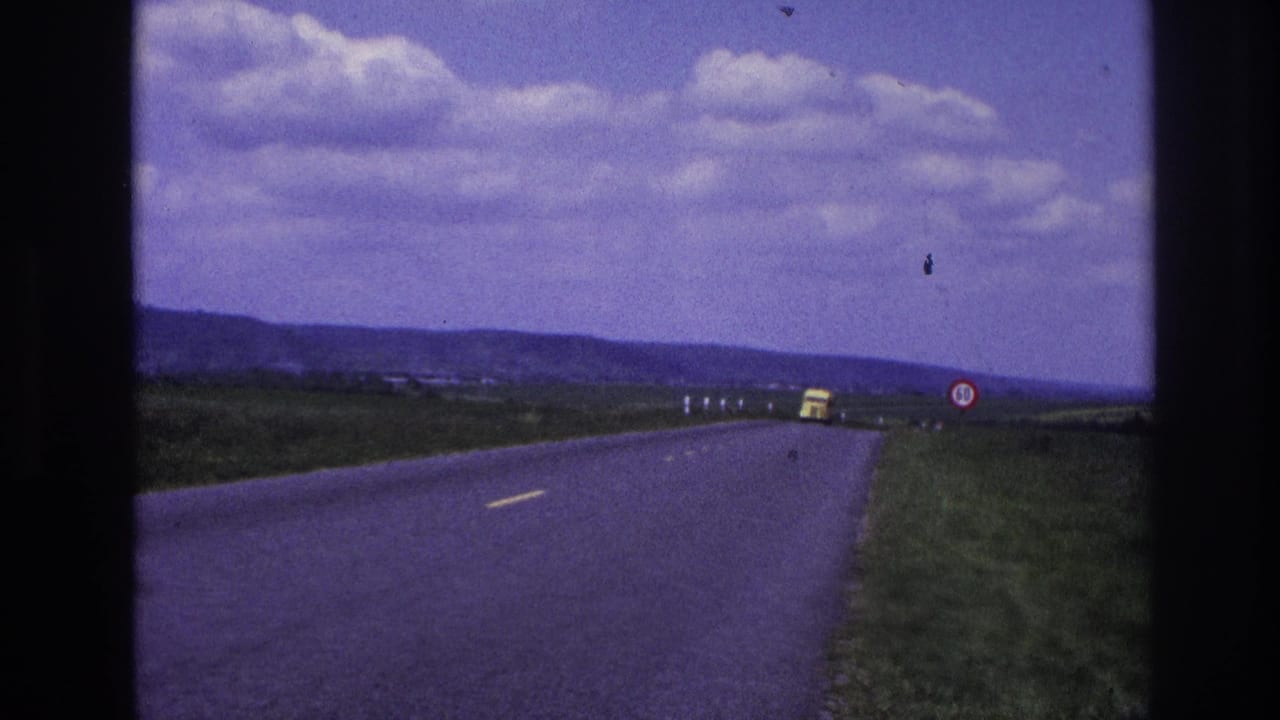 Two-lane rural road with a distant yellow truck, grassy fields, low hills, and clouded sky.