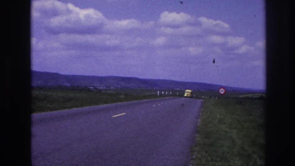 1967 France 8mm frame showing a distant yellow truck ahead on an open two-lane road through fields below low hills and scattered clouds.