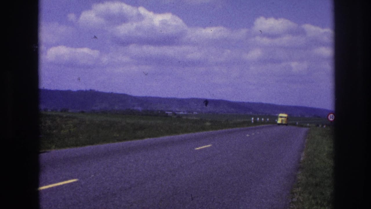 Open road curving through countryside with a yellow truck far ahead and a small roadside sign.