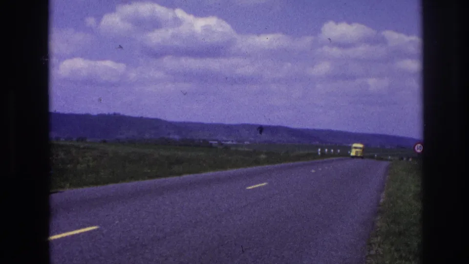 1967 France home-movie frame from a moving vehicle, with a yellow truck in the distance on a rural asphalt road bordered by fields and hills.