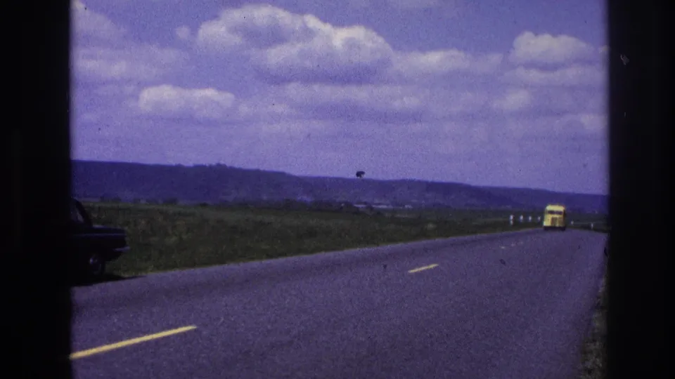 1967 France 8mm frame showing a moving roadside view with part of a dark car at the left edge and a yellow truck ahead on the rural road.