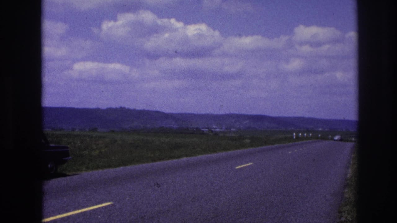 France 1967 8mm frame of a rural asphalt road under scattered clouds, with open fields, distant hills, and the edge of a dark stopped sedan entering at left.