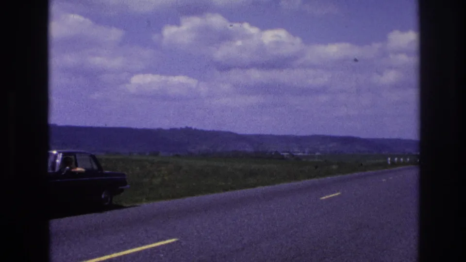 France 1967 8mm frame showing a dark sedan stopped on the roadside, with a person visible at the side window against fields, hills, and a cloudy sky.