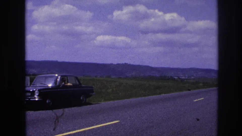 France 1967 8mm frame of a dark sedan near the roadside, partly cropped at left, with open fields, low hills, and a bright clouded sky behind it.