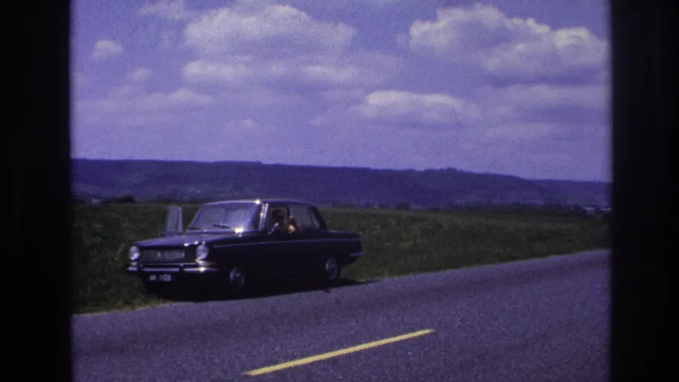 France 1967 8mm frame showing the stopped dark sedan more clearly on the grassy shoulder, with a person visible near the window and open countryside beyond.