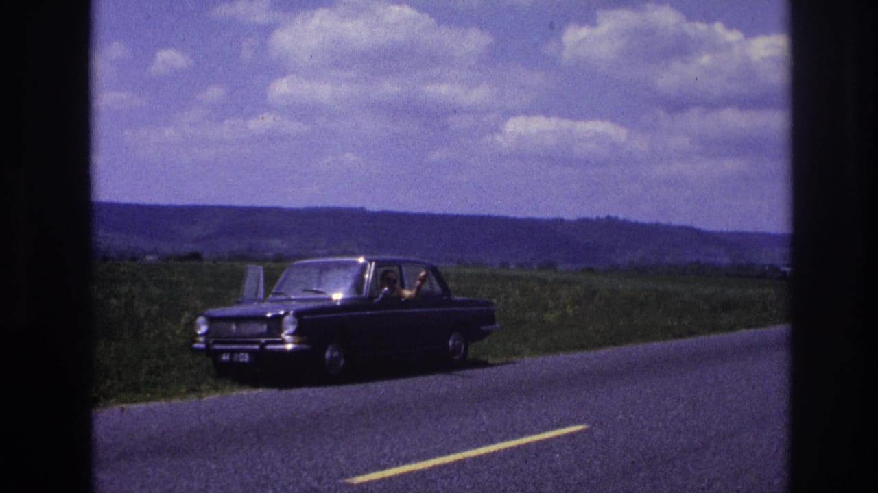 Dark European sedan on a rural two-lane road, person visible at the wheel, rolling green hills and cloudy sky behind.