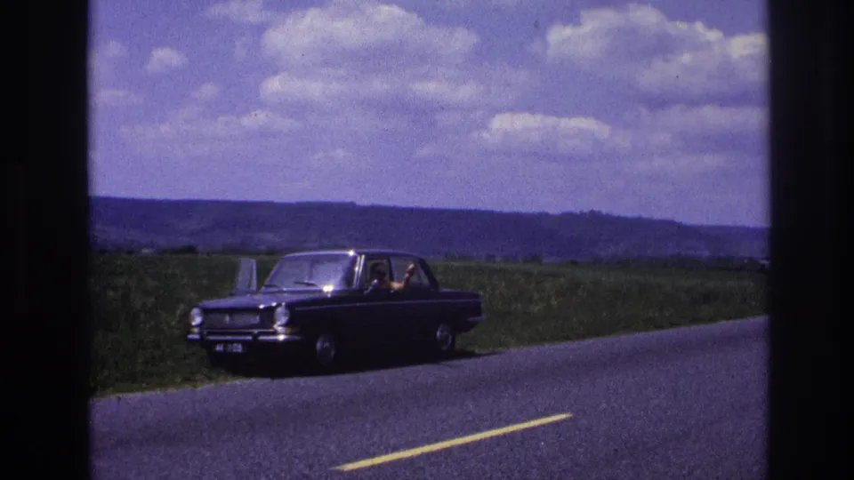 A dark 1960s European sedan on a rural French road, circa 1967; a figure is visible through the driver-side window against rolling open countryside.