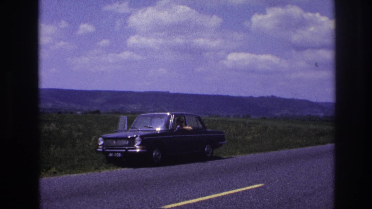 Dark 1960s European sedan on a rural French road, circa 1967; a figure is visible through the side windows with green fields and cumulus clouds in the background.