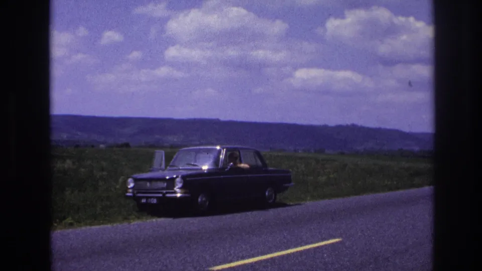 Dark 1960s European sedan on a rural French road, circa 1967; a figure is visible through the side windows with green fields and cumulus clouds in the background.
