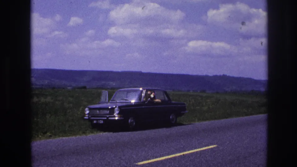 A figure waves from inside a dark 1960s European sedan stopped on a rural French road, circa 1967, with green fields and a cloudy sky visible behind.