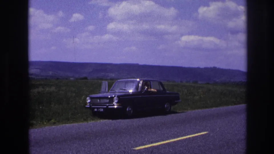 Dark European sedan on a rural French road, circa 1967; figure visible at the wheel with pastoral countryside and scattered clouds behind.