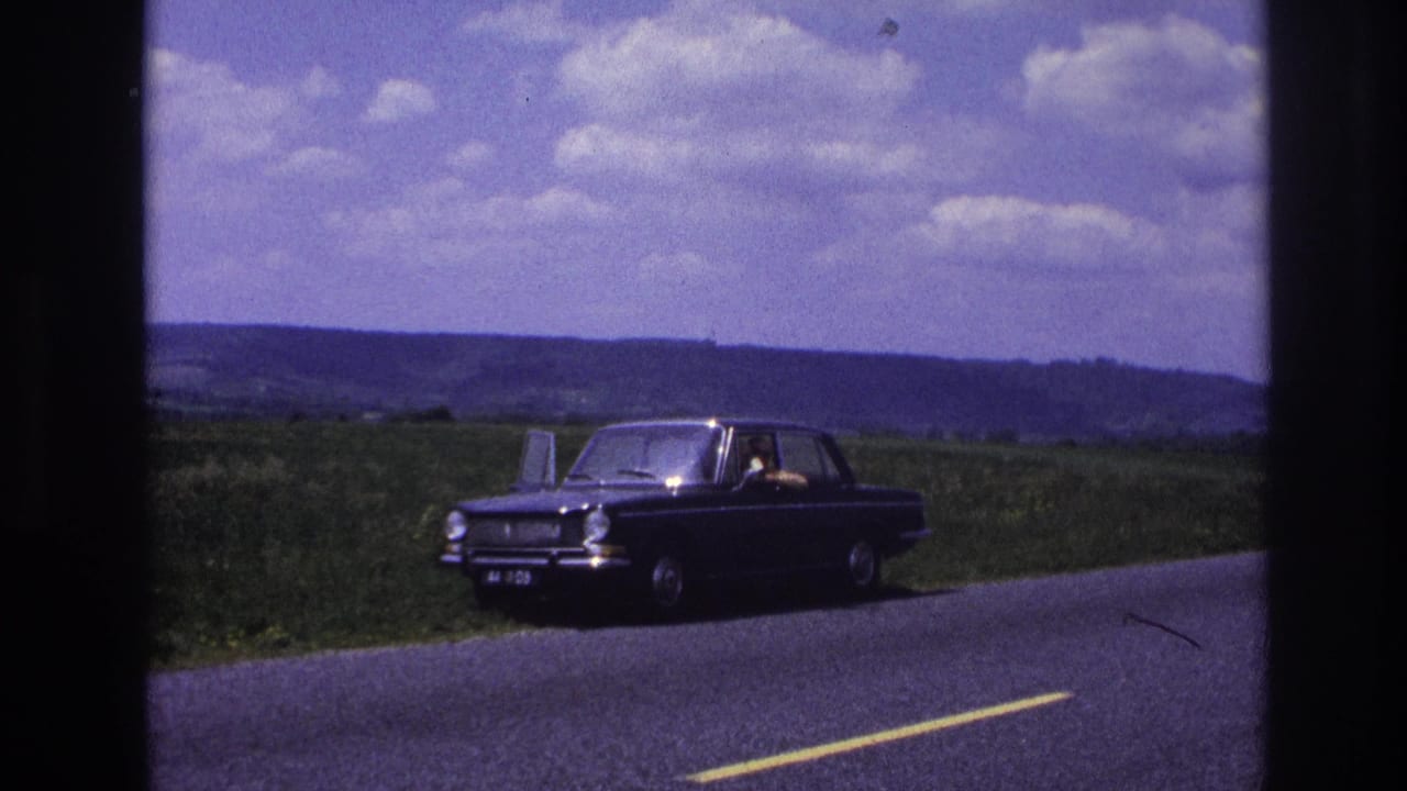 Dark compact sedan parked on a rural two-lane road, person visible inside, rolling green hills under a cloudy sky.