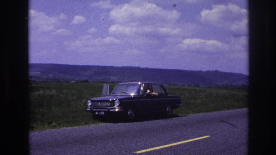 Dark compact European sedan parked on a country road in rural France, c. 1967; person visible in driver's seat, rolling green hills and cumulus clouds behind.
