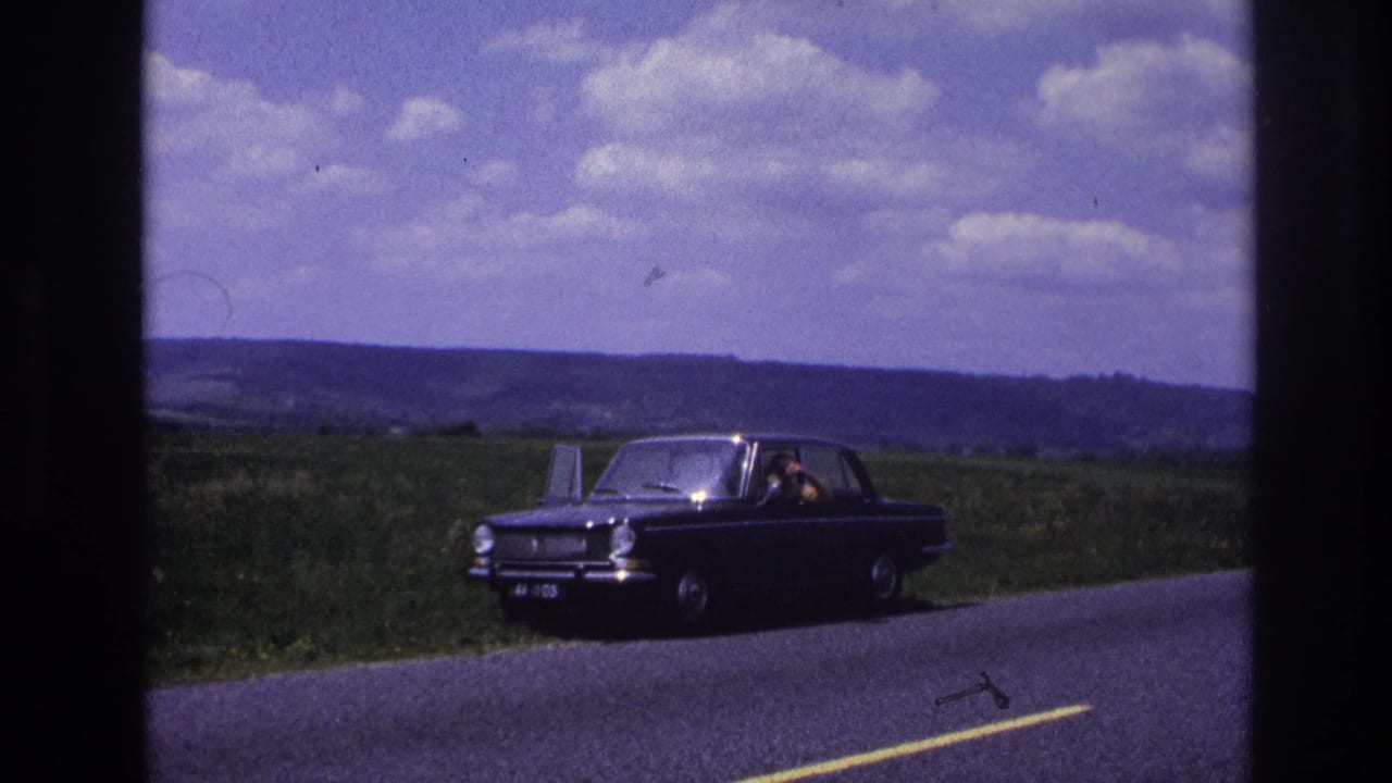 Dark compact sedan on a rural road in France, c. 1967; figure visible inside, green countryside and scattered clouds in background.