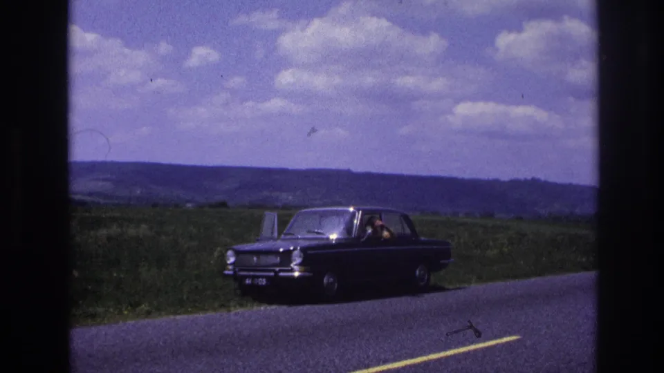 Dark compact sedan on a rural road in France, c. 1967; figure visible inside, green countryside and scattered clouds in background.