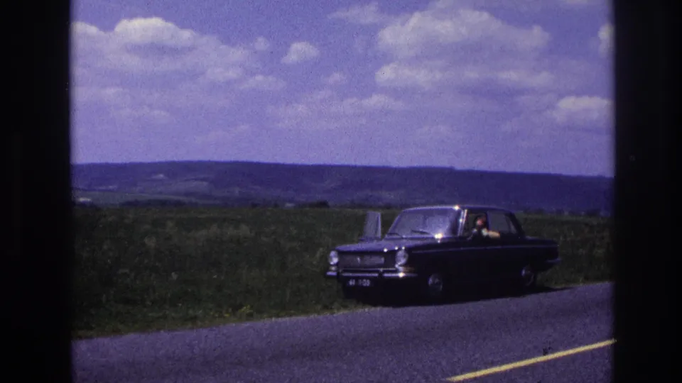 Dark European sedan parked on a rural French road, c. 1967, with driver-side door open; figure seated inside, rolling countryside and cumulus clouds behind.