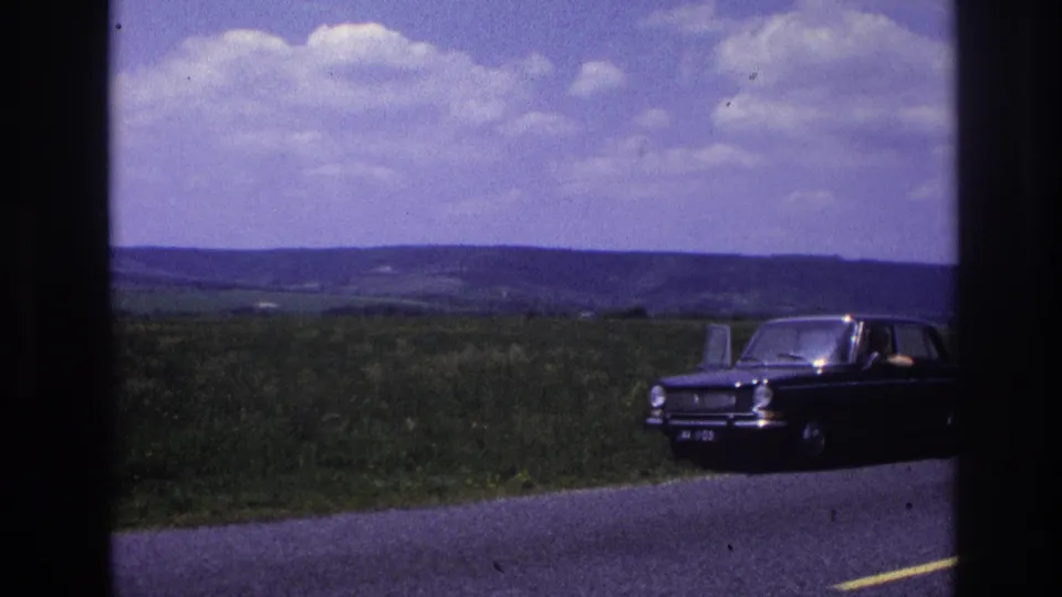 Dark compact European sedan parked at the edge of a rural French road, c. 1967; person visible inside, open door, green countryside and clouds in background.