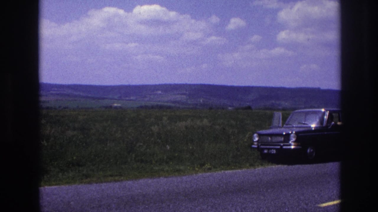 A dark-colored 1960s European sedan rests at the roadside in what appears to be rural France, 1967, open green fields and a wide sky stretching behind it.