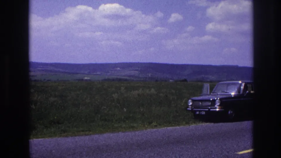 A dark-colored 1960s European sedan rests at the roadside in what appears to be rural France, 1967, open green fields and a wide sky stretching behind it.