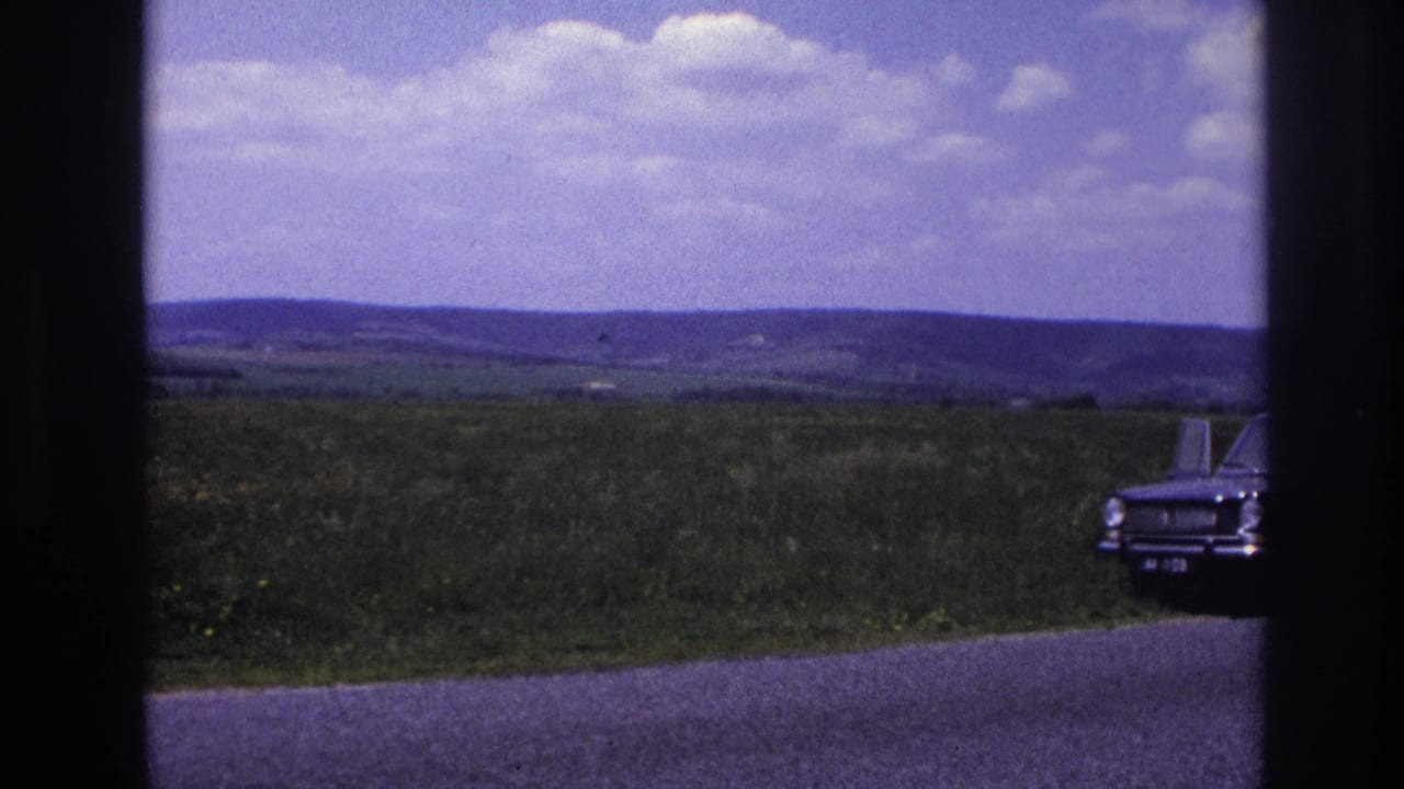 The same dark classic sedan seen from a slightly shifted angle along a rural French road, mid-1960s, open farmland and cumulus clouds filling the upper frame.
