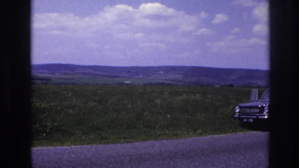 The same dark classic sedan seen from a slightly shifted angle along a rural French road, mid-1960s, open farmland and cumulus clouds filling the upper frame.