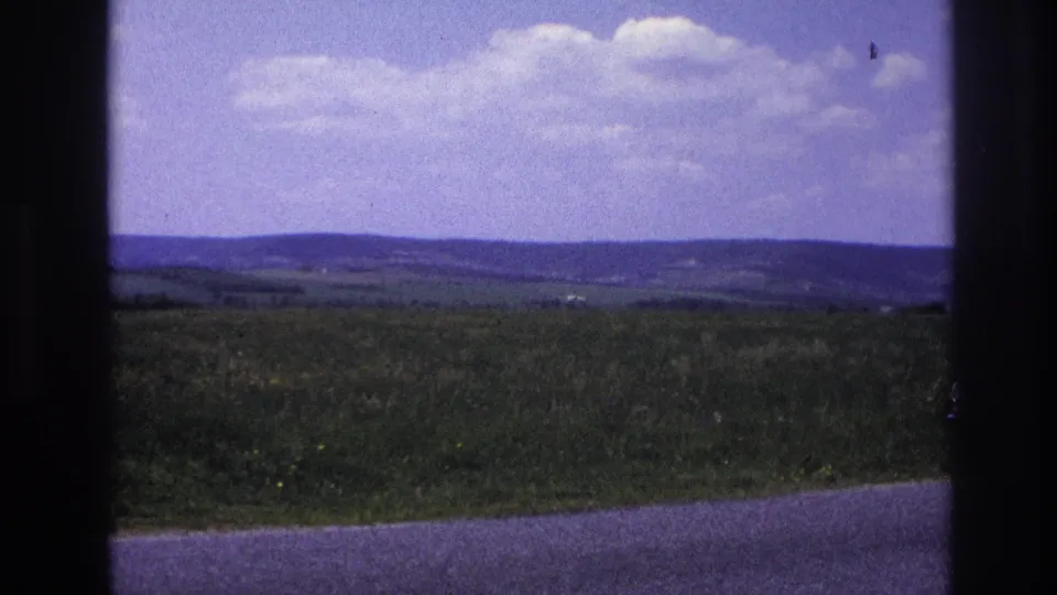 Wide open rural landscape, likely rural France c. 1967, with layered green hills receding to a distant horizon beneath a bright sky and road at the lower edge.
