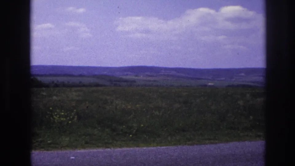 Pastoral French countryside, c. 1967, showing gently rolling agricultural land under a bright partly cloudy sky, shot on 8mm from a moving vehicle.