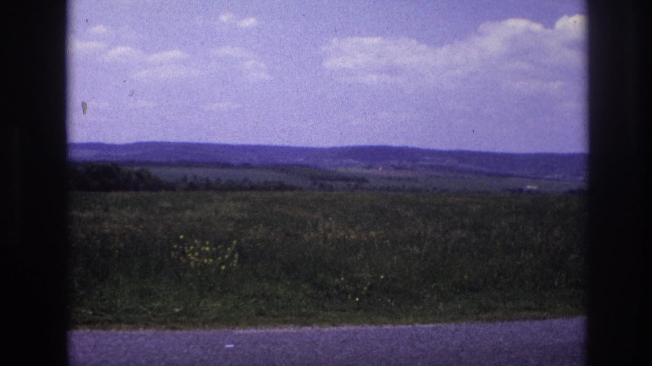 8mm home movie, France c.1967 — wide rural landscape with rolling green fields, distant wooded hills, and a large purple-tinged sky with scattered white clouds.